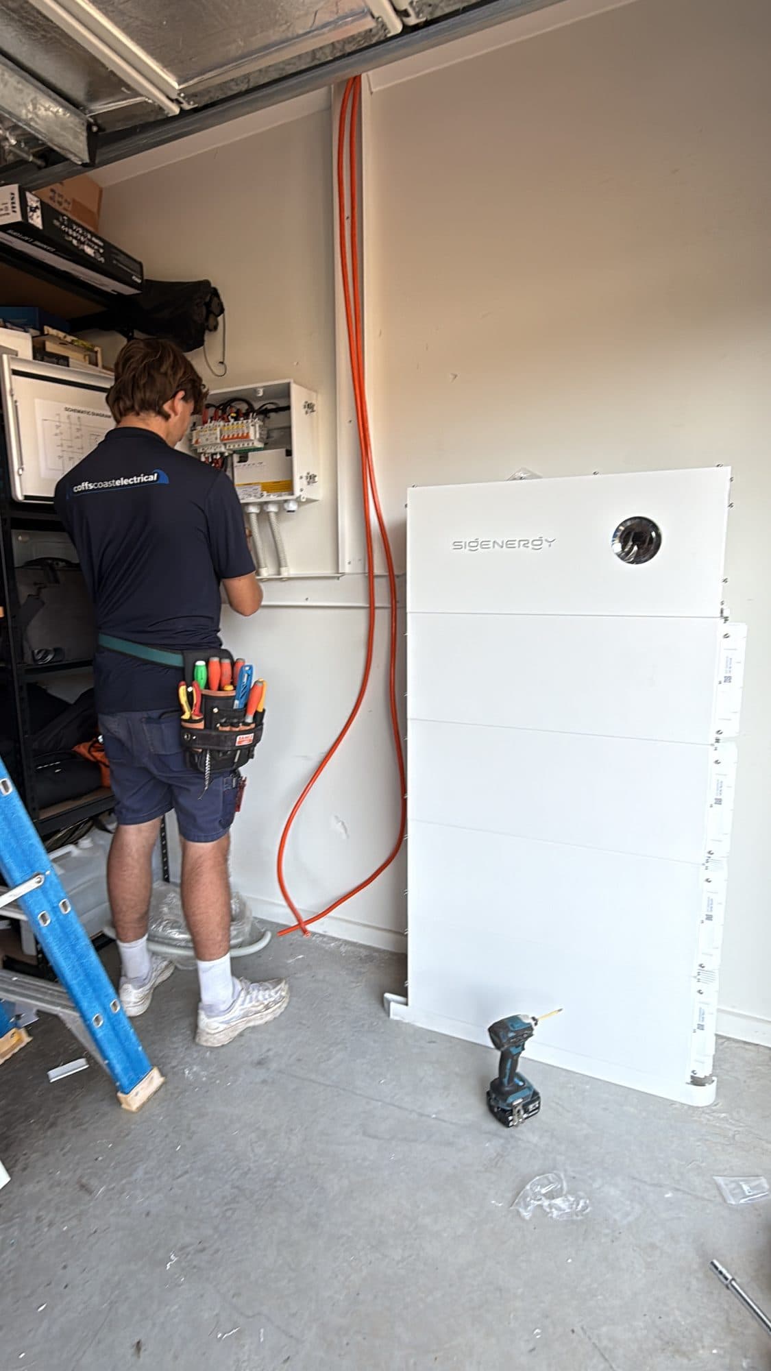 Sigenergy battery stack in a garage, installer wiring the switchboard beside it